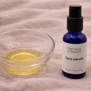 May include: A blue glass bottle of harvest & bloom face serum with a black pump dispenser and a white label reading Net Wt. 1 fl oz (30 g), positioned next to a clear glass bowl containing a small amount of golden yellow liquid oil.