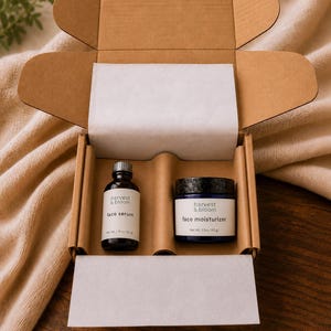 May include: A brown cardboard gift box containing a small glass bottle of Harvest & Bloom face serum with a 1 fl oz (30 g) label and a dark blue jar of Harvest & Bloom face moisturizer with a 1.5 oz (43 g) label, both nestled in a custom insert.