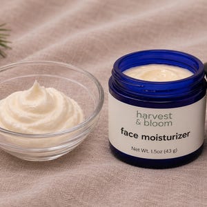 May include: A blue glass jar of Harvest & Bloom face moisturizer with a black lid, containing 1.5oz (43g) of cream, next to a clear glass bowl filled with a swirl of the same white face cream, showcasing the product texture and branding.