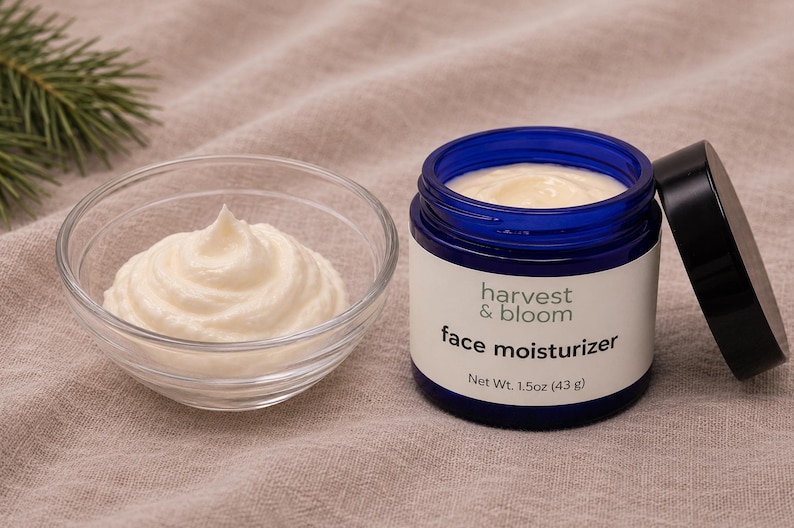 May include: A blue glass jar of Harvest & Bloom face moisturizer with a black lid, containing 1.5oz (43g) of cream, next to a clear glass bowl filled with a swirl of the same white face cream, showcasing the product texture and branding.