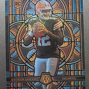 May include: A football trading card featuring Shedeur Sanders of the Browns. The card has a stained glass design with a mosaic pattern. The player is wearing a brown helmet and uniform, holding a football. The card is labeled "Rookie Card".