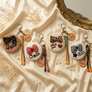 Pode incluir: Uma coleção de chaveiros fofos com vários designs. Cada chaveiro apresenta uma base redonda e felpuda com um laço ou um coração. Alguns têm etiquetas de madeira com nomes como "Olivia" e "Isabella". Os chaveiros têm argolas de metal e borlas coloridas.