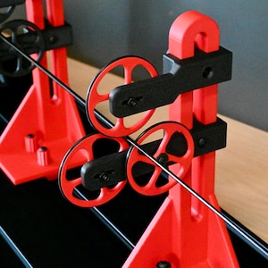 May include: A mechanical filament guide system featuring two red vertical support towers mounted on a black base. Each tower holds two black horizontal arms with red circular wheels. A thin black filament thread runs through the wheels.