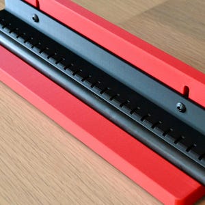 May include: A red L-shaped bracket with a black metal insert featuring a series of evenly spaced vertical slots. A thin black rod is positioned horizontally within the channel of the bracket, which is secured by two visible black screws.