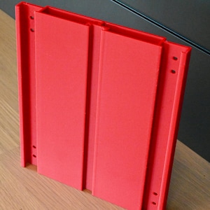 May include: A bright red, rectangular mounting bracket featuring a central vertical channel and two side panels. The structure includes four small circular holes on the outer edges, designed for attachment, with a matte finish and uniform color.