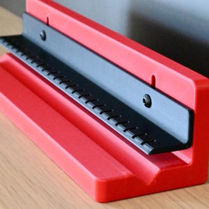May include: A red L-shaped stand featuring a black L-bracket attached to the vertical back panel. The black bracket has a series of evenly spaced vertical notches along its top edge. The stand has a grooved base for holding items upright.