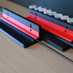 May include: A black and red fishing rod holder with a rod in place. The holder has a black base and a red top with angled slots. Spools of thread in various colors are visible in the background.