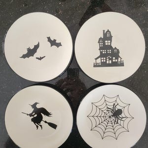May include: Set of four white ceramic plates with black Halloween-themed designs. Includes bats, a haunted house, a witch on a broom, and a spiderweb with a spider. Each plate has a black rim.