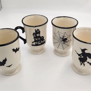 May include: Four cream-colored Halloween mugs with black accents and handles. Each mug features a different black graphic: bats, a haunted house, a spiderweb with a spider, and a witch on a broomstick. The mugs have a pedestal base.