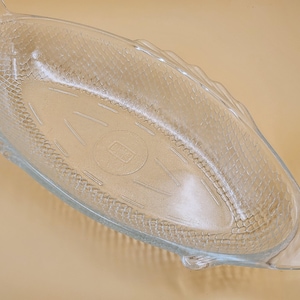 Peut inclure: Un plat de service en verre transparent en forme de poisson. Le plat présente une surface texturée ressemblant à des écailles de poisson et des détails de nageoires et de queue. L'intérieur comporte des lignes gravées et un emblème circulaire.