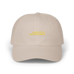 Puede incluir: Gorra de béisbol beige con visera curva. La gorra presenta el texto bordado "LIGHT IT UP LIKE DYNAMITE" en hilo amarillo en la parte delantera. El sombrero es un diseño clásico de seis paneles con una correa ajustable en la parte posterior.