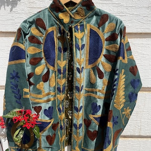 May include: A sage green velvet jacket featuring intricate embroidery in shades of gold, dark blue, and brown. The design includes stylised floral, leaf, and heart-shaped motifs across the front and collar, with a visible patterned inner lining.