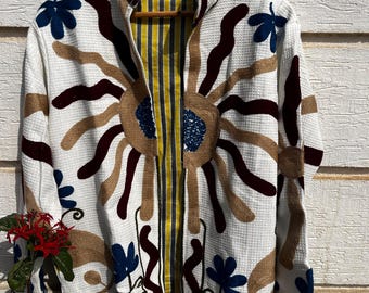 May include: A white waffle-textured jacket featuring large embroidered sun motifs in tan and dark maroon, accented with small blue floral patterns. The interior lining displays a vertical yellow, white, and grey striped fabric pattern.