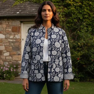 May include: A navy blue quilted jacket featuring a white floral pattern, a mandarin collar, and long sleeves with folded cuffs. The jacket has two front patch pockets and is worn over a white top and dark blue denim jeans.