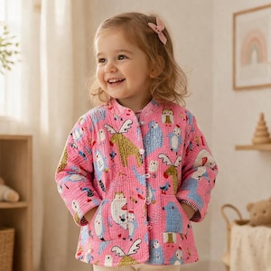 May include: A young child wearing a pink quilted jacket with a whimsical pattern of reindeer and other creatures. The jacket has a button-up front and is paired with cream-colored leggings and matching shoes. The child is smiling.