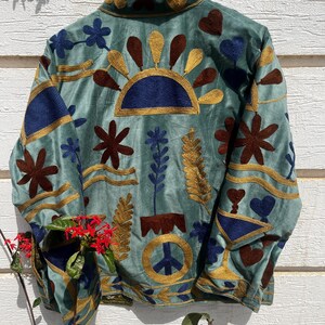 Puede incluir: Una chaqueta verde salvia con bordados intrincados en tonos azul oscuro, marr&oacute;n y dorado. El dise&ntilde;o incluye un s&iacute;mbolo de paz central, motivos florales, formas de coraz&oacute;n y patrones solares. La chaqueta tiene cuello alto y mangas largas.