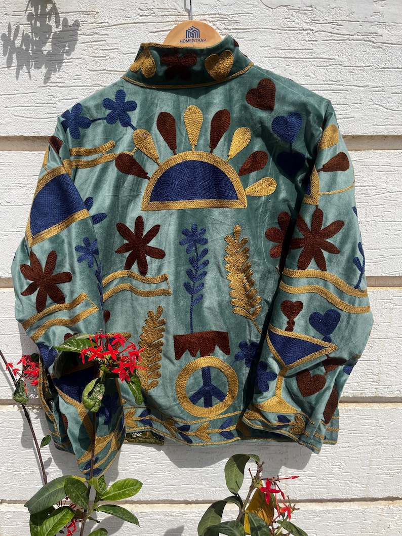 Puede incluir: Una chaqueta verde salvia con bordados intrincados en tonos azul oscuro, marr&oacute;n y dorado. El dise&ntilde;o incluye un s&iacute;mbolo de paz central, motivos florales, formas de coraz&oacute;n y patrones solares. La chaqueta tiene cuello alto y mangas largas.