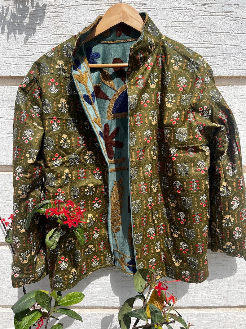 Puede incluir: Una chaqueta reversible con exterior verde oliva y un patr&oacute;n floral repetitivo en rojo, blanco y amarillo. El forro interior es de tela azul claro adornada con grandes y llamativos motivos florales y de hojas bordados en azul oscuro, marr&oacute;n y dorado.