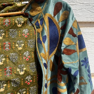 Puede incluir: Una chaqueta azul claro con bordados intrincados en tonos azul oscuro, marr&oacute;n y dorado. El forro interior es de tela verde oliva con un patr&oacute;n floral repetitivo en blanco, rojo y amarillo. La prenda cuelga de una percha de madera.