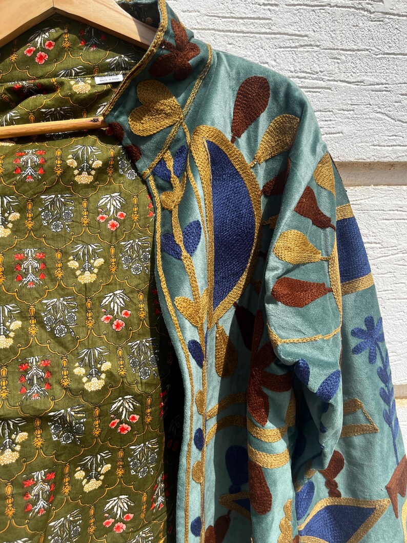 Puede incluir: Una chaqueta azul claro con bordados intrincados en tonos azul oscuro, marr&oacute;n y dorado. El forro interior es de tela verde oliva con un patr&oacute;n floral repetitivo en blanco, rojo y amarillo. La prenda cuelga de una percha de madera.