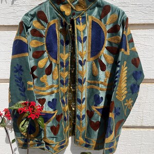 Puede incluir: Una chaqueta de terciopelo verde salvia con intrincados bordados en tonos dorados, azul oscuro y marr&oacute;n. El dise&ntilde;o incluye patrones de rayos de sol estilizados, formas de coraz&oacute;n y motivos de hojas. La chaqueta tiene cuello alto y apertura frontal.