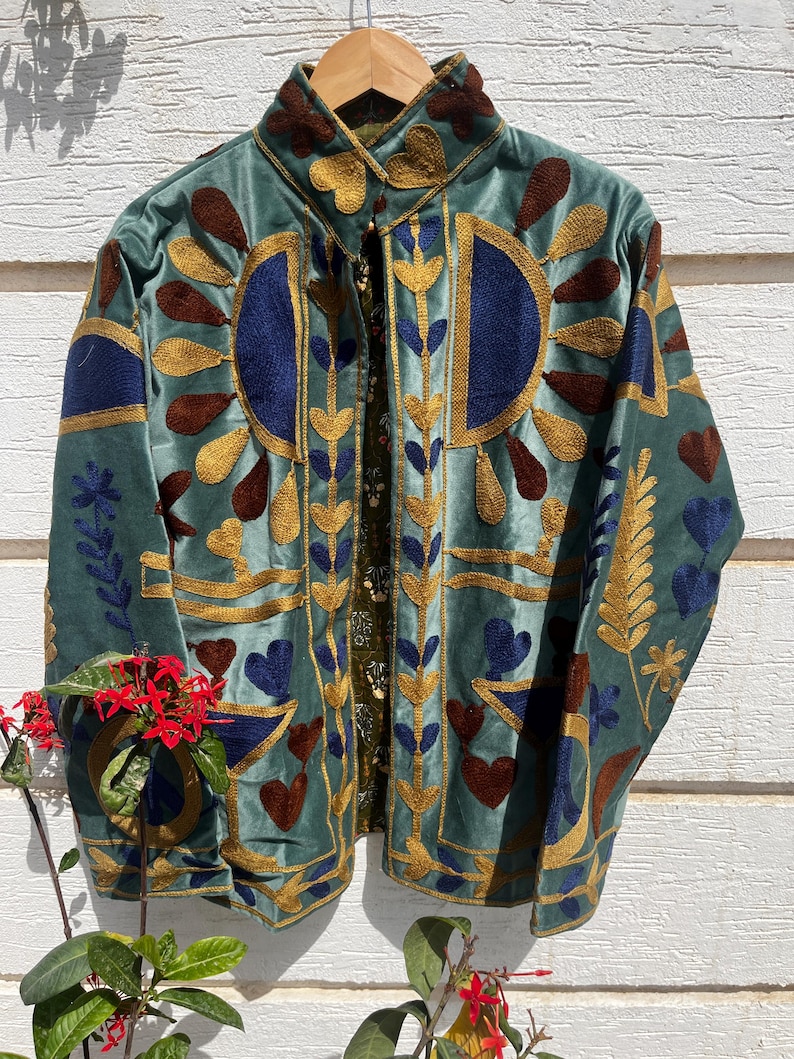 Puede incluir: Una chaqueta de terciopelo verde salvia con intrincados bordados en tonos dorados, azul oscuro y marr&oacute;n. El dise&ntilde;o incluye patrones de rayos de sol estilizados, formas de coraz&oacute;n y motivos de hojas. La chaqueta tiene cuello alto y apertura frontal.