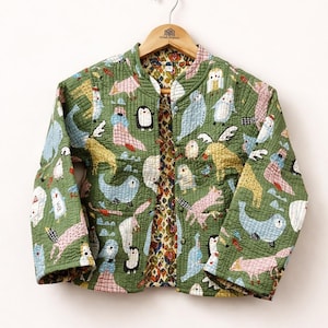 Puede incluir: Chaqueta acolchada verde con un estampado caprichoso de pájaros y animales. La chaqueta tiene cuello redondo y mangas largas. El forro interior presenta un estampado floral. La chaqueta está colgada de una percha de madera.