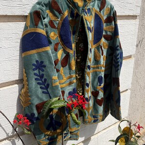 Puede incluir: Una chaqueta azul claro con intrincados bordados en dorado, azul oscuro y marr&oacute;n. El dise&ntilde;o incluye motivos florales, formas geom&eacute;tricas y un s&iacute;mbolo de la paz. La chaqueta tiene un cuello alto y un forro interior oscuro con estampado floral.
