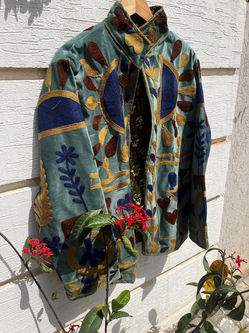Puede incluir: Una chaqueta azul claro con intrincados bordados en dorado, azul oscuro y marr&oacute;n. El dise&ntilde;o incluye motivos florales, formas geom&eacute;tricas y un s&iacute;mbolo de la paz. La chaqueta tiene un cuello alto y un forro interior oscuro con estampado floral.