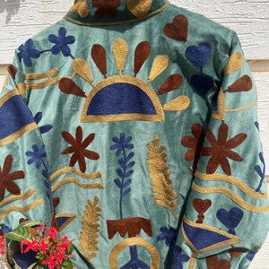 Puede incluir: Una chaqueta de color verde claro con bordados intrincados en tonos azul oscuro, marr&oacute;n y dorado. El dise&ntilde;o incluye motivos florales, formas de coraz&oacute;n, un sol y un s&iacute;mbolo de la paz. La chaqueta cuelga de una percha de madera con un logotipo.