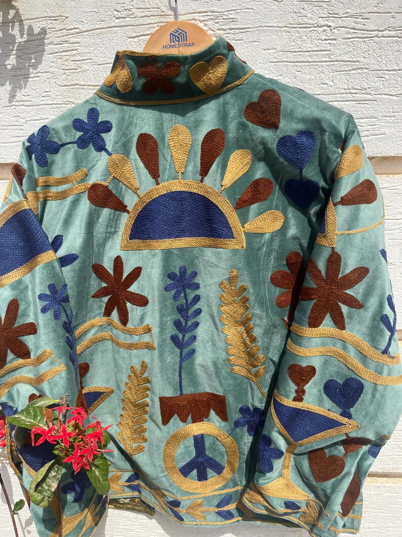 Puede incluir: Una chaqueta de color verde claro con bordados intrincados en tonos azul oscuro, marr&oacute;n y dorado. El dise&ntilde;o incluye motivos florales, formas de coraz&oacute;n, un sol y un s&iacute;mbolo de la paz. La chaqueta cuelga de una percha de madera con un logotipo.