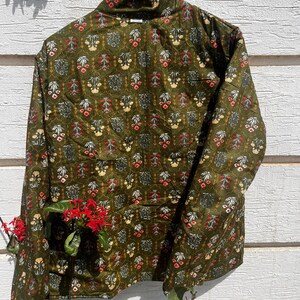 Puede incluir: Una chaqueta acolchada de manga larga en color verde oliva con un patr&oacute;n floral repetitivo en tonos rojo, blanco y amarillo. La chaqueta tiene cuello alto, cierre frontal de botones y un corte estructurado sobre una percha de madera.