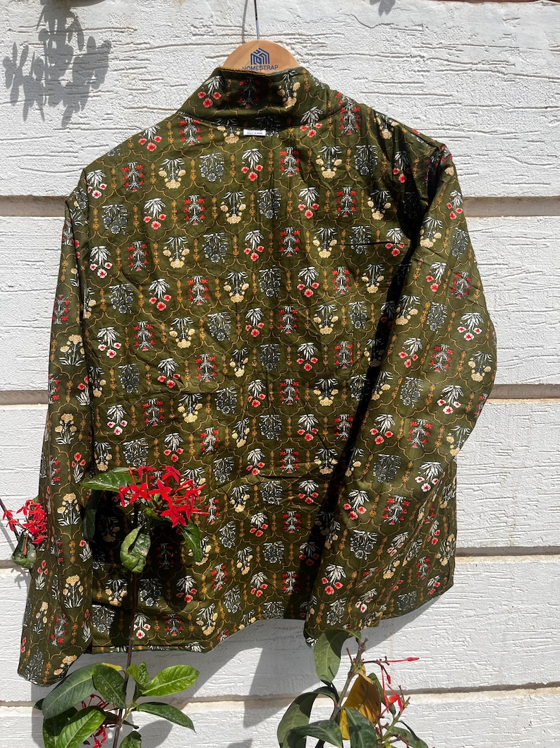 Puede incluir: Una chaqueta acolchada de manga larga en color verde oliva con un patr&oacute;n floral repetitivo en tonos rojo, blanco y amarillo. La chaqueta tiene cuello alto, cierre frontal de botones y un corte estructurado sobre una percha de madera.