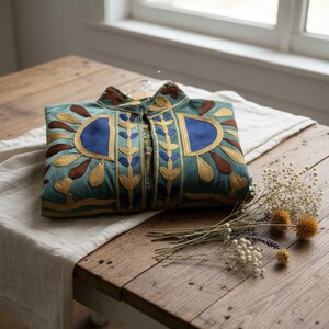 Puede incluir: Una chaqueta de color verde azulado con intrincados bordados en tonos dorados, marrones y azules. El dise&ntilde;o incluye parches azules semicirculares en el pecho, patrones florales verticales y un cuello alto, todo en una disposici&oacute;n plegada.