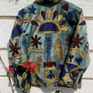 Puede incluir: Una chaqueta verde salvia con bordados intrincados en tonos azul oscuro, marr&oacute;n y dorado. El dise&ntilde;o incluye motivos florales, formas de coraz&oacute;n, un semic&iacute;rculo similar a un sol y un s&iacute;mbolo de la paz. La chaqueta tiene cuello alto y mangas largas.