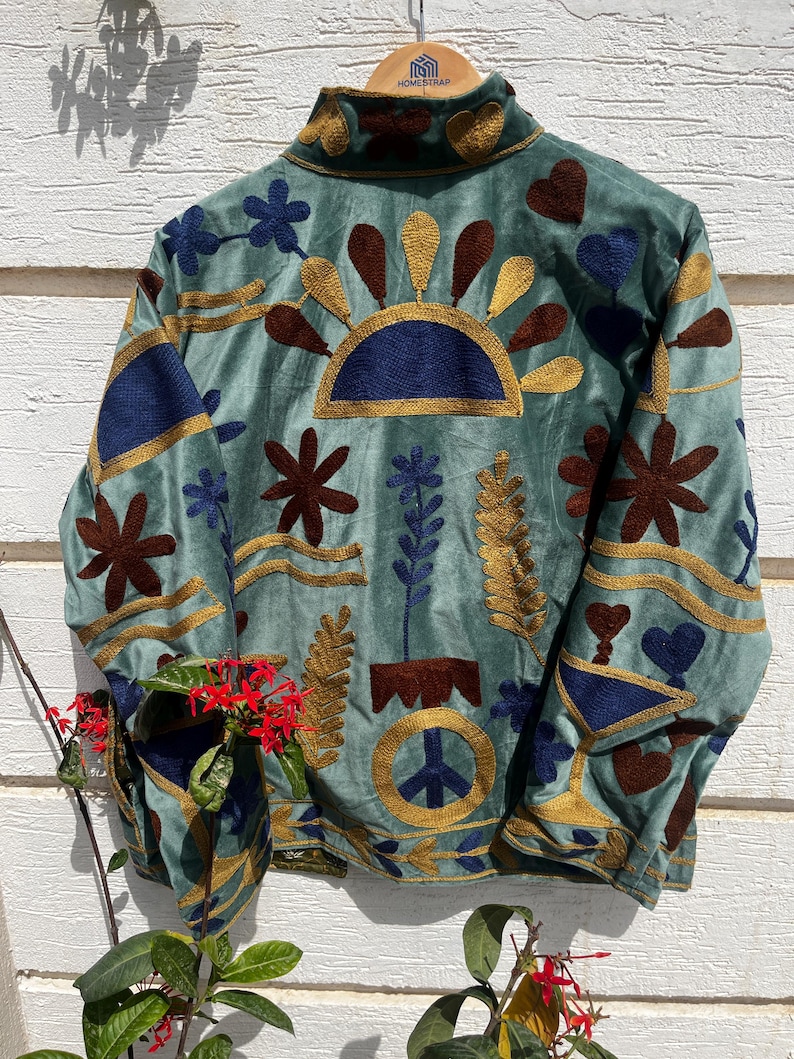 Puede incluir: Una chaqueta verde salvia con bordados intrincados en tonos azul oscuro, marr&oacute;n y dorado. El dise&ntilde;o incluye motivos florales, formas de coraz&oacute;n, un semic&iacute;rculo similar a un sol y un s&iacute;mbolo de la paz. La chaqueta tiene cuello alto y mangas largas.