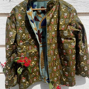Puede incluir: Una chaqueta acolchada con exterior verde oliva y un patr&oacute;n floral repetitivo en rojo, blanco y amarillo. El forro interior es de tela azul claro adornada con grandes motivos florales y de hojas bordados en tonos oscuros.