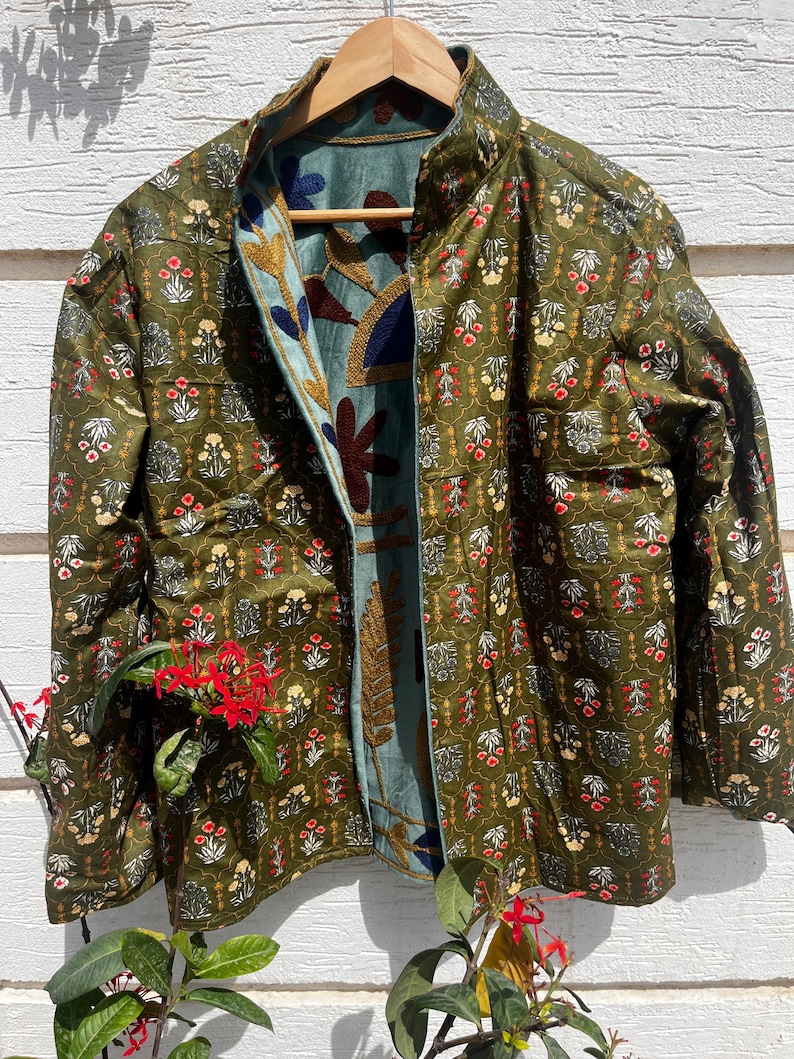 Puede incluir: Una chaqueta acolchada con exterior verde oliva y un patr&oacute;n floral repetitivo en rojo, blanco y amarillo. El forro interior es de tela azul claro adornada con grandes motivos florales y de hojas bordados en tonos oscuros.