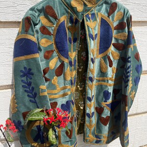 Puede incluir: Una chaqueta de terciopelo azul claro con bordados intrincados en dorado, azul oscuro y marr&oacute;n. El dise&ntilde;o incluye patrones florales y geom&eacute;tricos en el frente y las mangas. La chaqueta tiene un cuello alto y un forro verde oscuro con estampado floral.