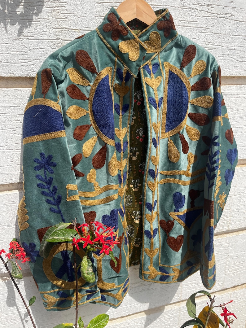 Puede incluir: Una chaqueta de terciopelo azul claro con bordados intrincados en dorado, azul oscuro y marr&oacute;n. El dise&ntilde;o incluye patrones florales y geom&eacute;tricos en el frente y las mangas. La chaqueta tiene un cuello alto y un forro verde oscuro con estampado floral.