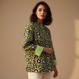 Op de afbeelding: Een groene gewatteerde jas met een zwart-roze luipaardprint. De jas heeft een opstaande kraag en groen-wit gestreepte manchetten. Het model draagt de jas met een crèmekleurige broek.