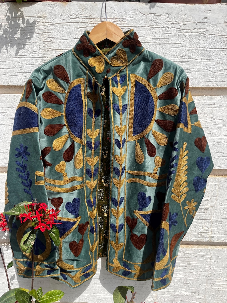 Puede incluir: Una chaqueta de terciopelo verde salvia con bordados intrincados en tonos dorados, azul oscuro y marr&oacute;n. El dise&ntilde;o incluye motivos estilizados de flores, hojas y corazones en la parte delantera y el cuello, con un forro interior estampado.