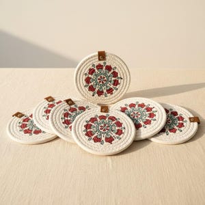 May include: A set of six circular coasters made from coiled off-white rope. Each coaster features a central mandala-style pattern in red and teal, with a small brown faux leather tab attached to the edge by a metallic rivet for hanging or storage.