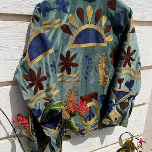 Puede incluir: Una chaqueta verde salvia con bordados intrincados en tonos azul oscuro, marr&oacute;n y dorado. El dise&ntilde;o incluye motivos florales, patrones de rayos de sol y un s&iacute;mbolo de la paz. La chaqueta tiene cuello alto y mangas largas con bordados a juego.