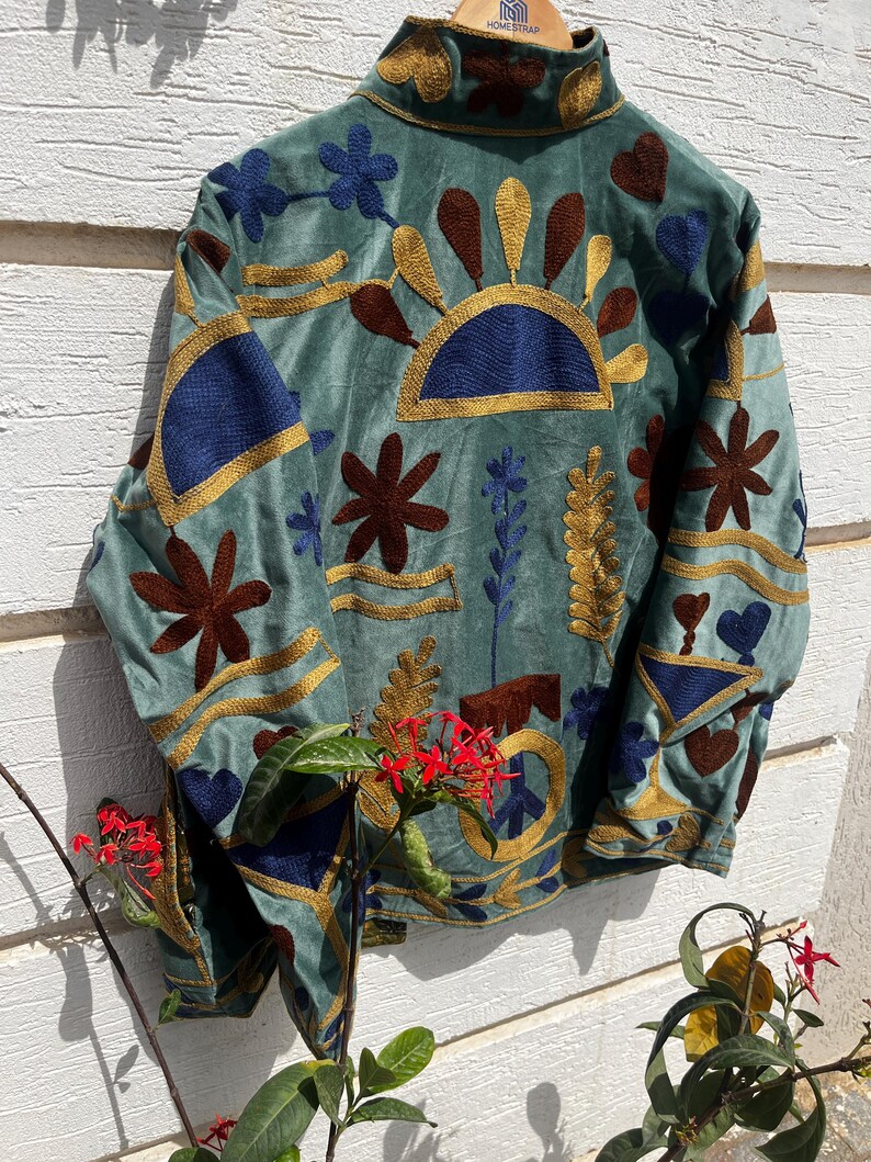 Puede incluir: Una chaqueta verde salvia con bordados intrincados en tonos azul oscuro, marr&oacute;n y dorado. El dise&ntilde;o incluye motivos florales, patrones de rayos de sol y un s&iacute;mbolo de la paz. La chaqueta tiene cuello alto y mangas largas con bordados a juego.