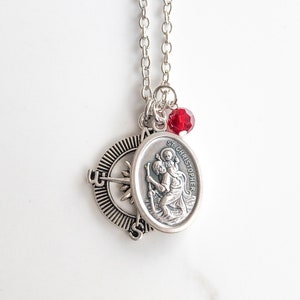 Op de afbeelding: Zilveren ketting met een zilveren kompasbedel, een zilveren ovale bedel met de tekst "St. Christopher" en een rode kristallen bedel.