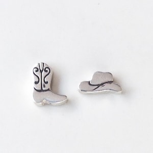 May include: A pair of small silver-toned metal charms consisting of one cowboy boot with engraved black line detailing on the shaft and one cowboy hat with a curved brim and a simple indented line representing the crown and band.