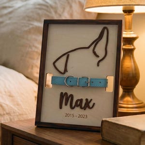 Puede incluir: Un marco de madera conmemorativo para mascotas con una silueta de perro, un collar azul claro y el nombre "Max" con las fechas 2015-2023. El marco está sobre una mesita de noche de madera junto a una lámpara y un libro.