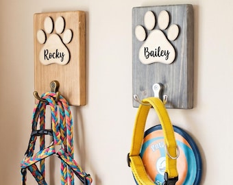 May include: Three rectangular wooden wall hooks with paw print cutouts and personalized names Milo, Rocky, and Bailey. Each plaque features a metal hook holding pet accessories, including a pink sweater, a colorful leash, and a yellow collar.