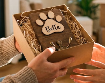 May include: A rectangular dark wood plaque inside a cardboard gift box filled with paper filler. The plaque features a light wood paw print cutout with the name Bella printed in black script and a brass-colored metal hook attached at the bottom.